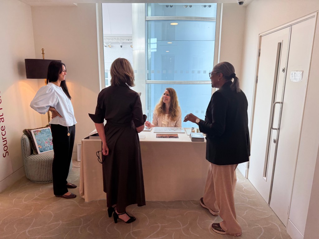 Eine Gruppe von vier Personen spricht anlässlich einer Maldemonstration an einem Tisch in einem eleganten Showroom während der London Craft Week, mit einem hellen Fenster des London Harbour Design Centres im Hintergrund