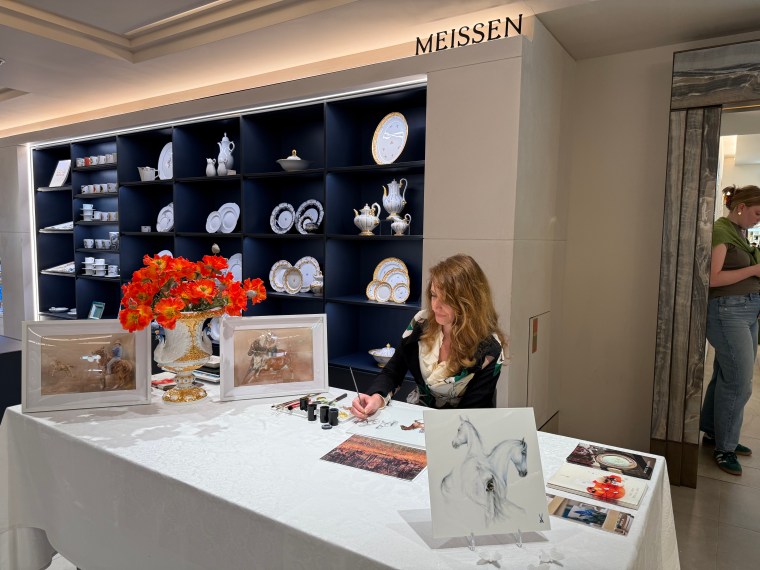Künstlerin Michaela Stocker arbeitet an einem Tisch und malt Pferde, umgeben von Porzellan und Kunstwerken, während im Hintergrund Geschirr und Dekorationsobjekte ausgestellt sind. Auf dem Tisch stehen Blumen und Malutensilien.