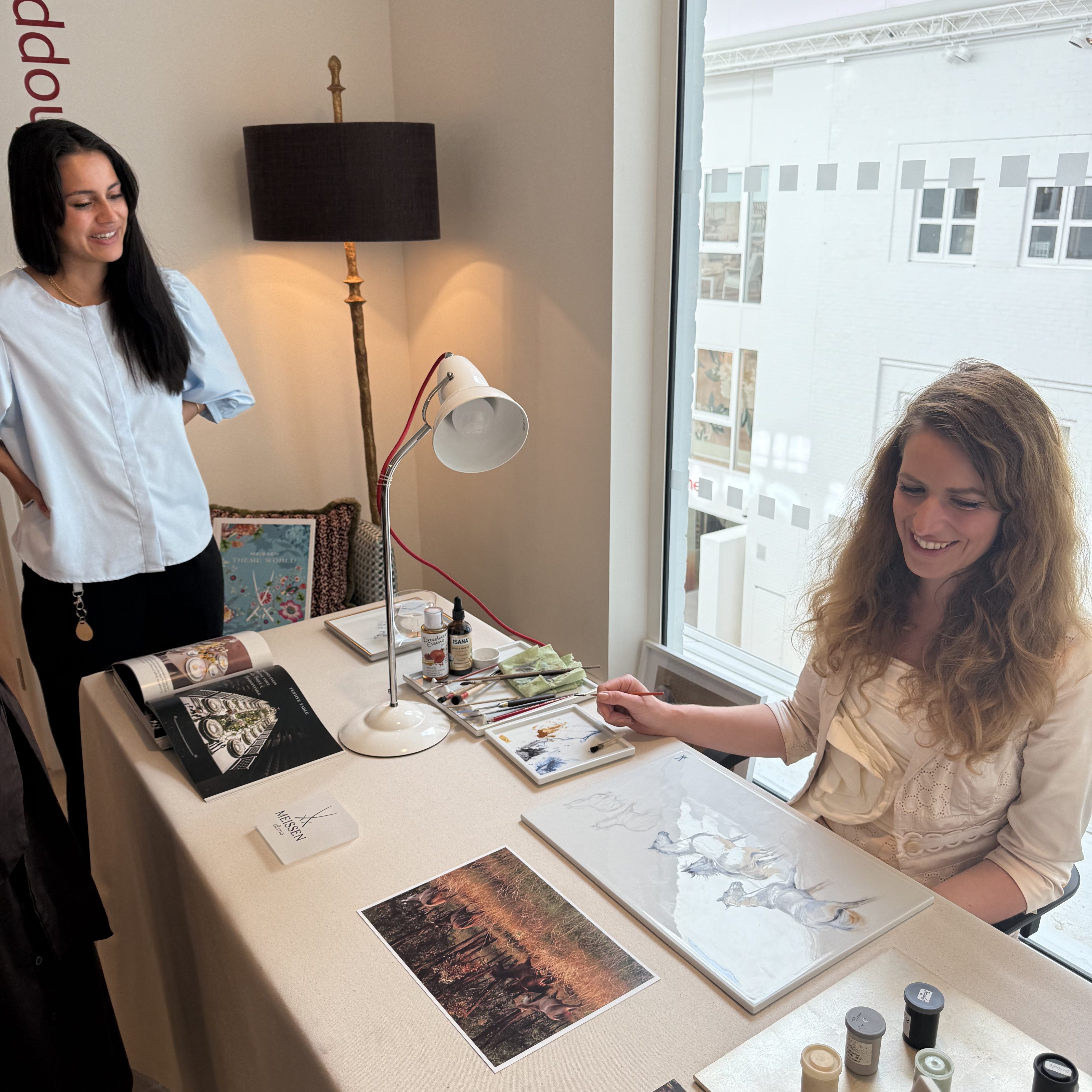 Zwei Frauen in einem hellen Raum in dem The Source Showroom des Chelsea Harbour Design Centres, Künstlerin Michaela Stocker sitzt an einem Tisch und malt Pferde auf ein Wandgemälde aus Porzellan, während die andere daneben steht.