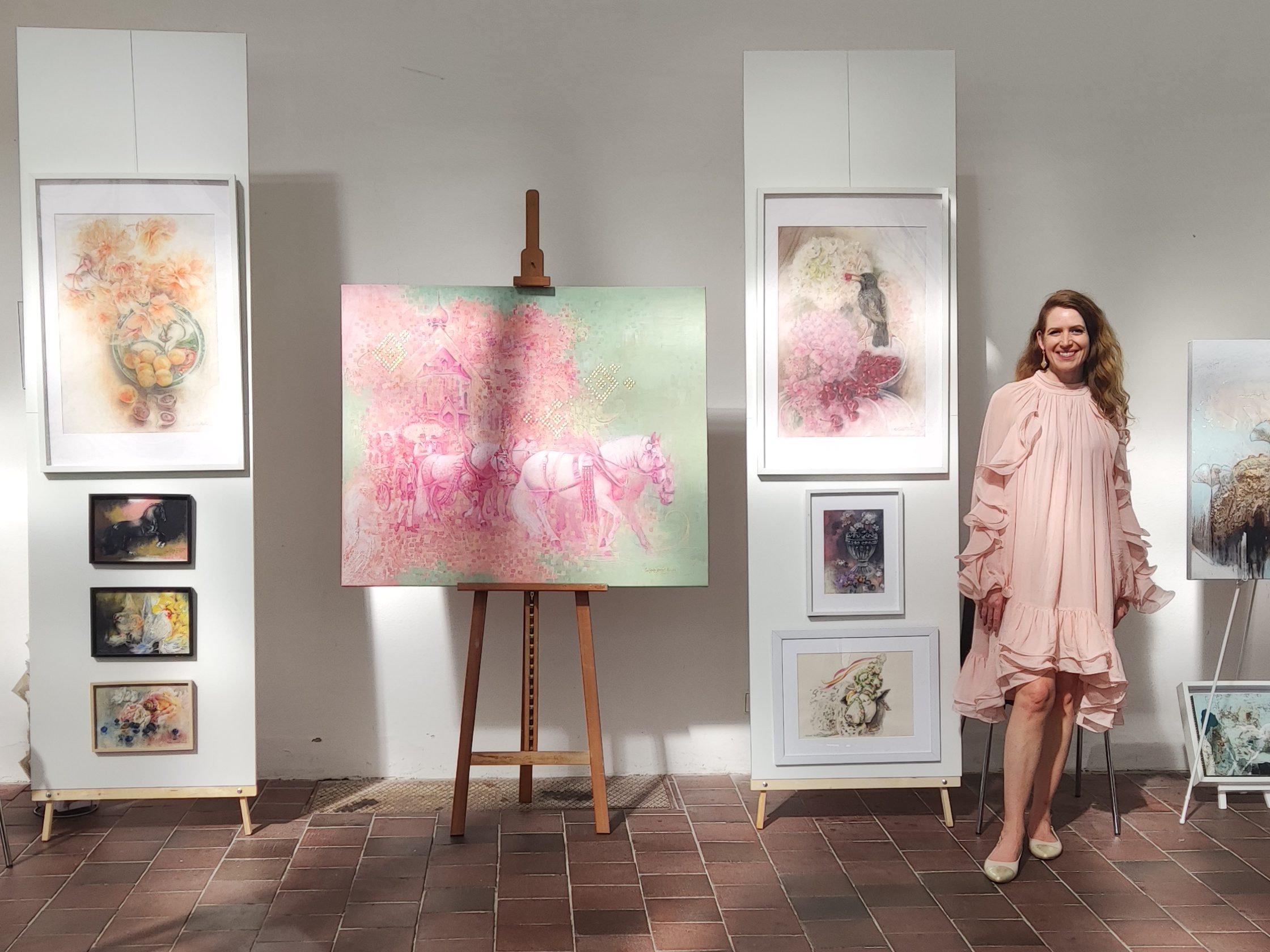 Michaela Stocker in einem hellrosa Kleid steht neben einer Galerie mit ihren Kunstwerken im Eisernen Haus im Schlosspark Nymphenburg München. Ausgestellt sind verschiedene Bilder in Rahmen dekoriert, darunter Pastellmalereien und impressionistische Motive in Öl.