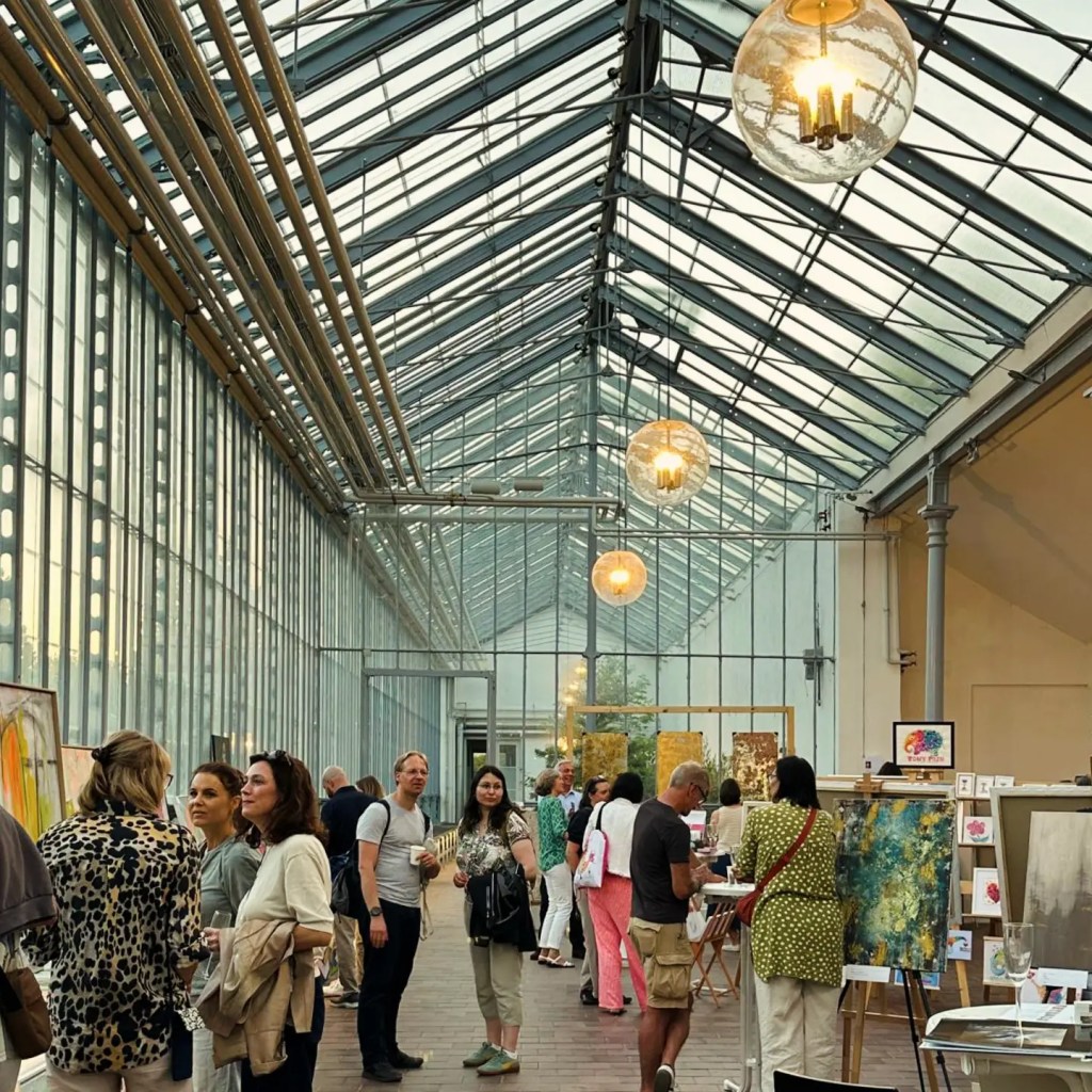 Ein kunstvolles Innenansicht des Eisernen Hauses im Schloss Nymphenburg während einer Ausstellung, mit Besuchern, die Bilder betrachten und sich unterhalten.